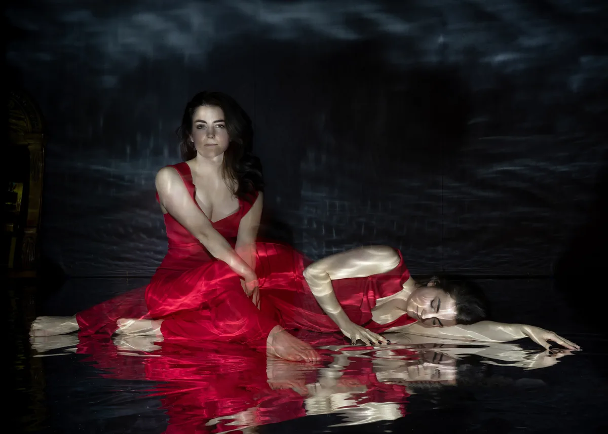 A woman in a flowing red dress appears twice, sitting upright with hands clasped and lying on her side with eyes closed, reflected on a dark, rippled surface with soft lighting.