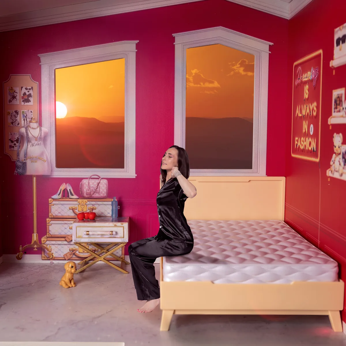 A woman in black satin pajamas sits barefoot on the edge of a cream-colored bed in a pink room with two windows showing an orange sunset over mountains.