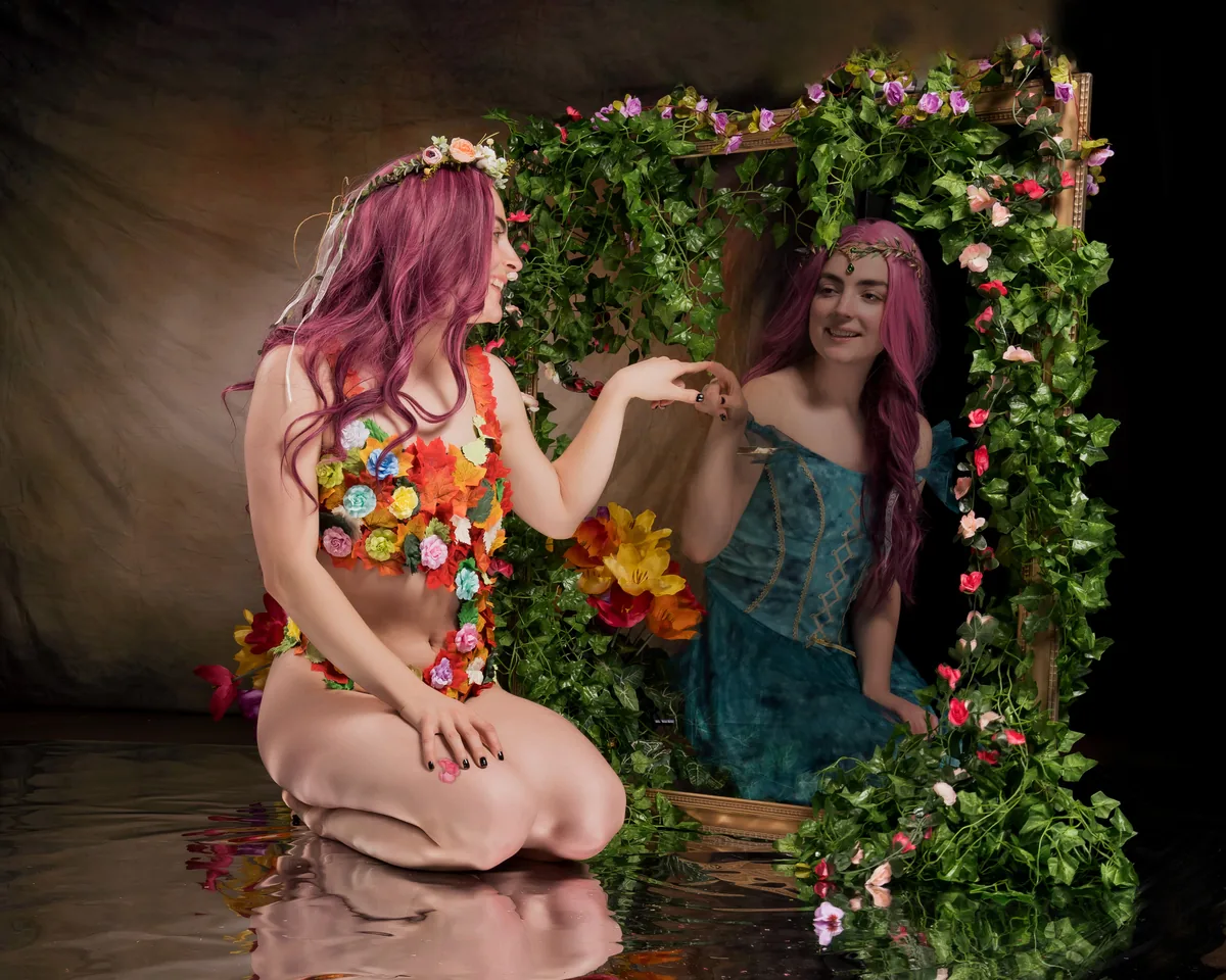 A woman with purple hair adorned with a floral wreath kneels beside a large mirror decorated with green ivy and pink flowers, her reflection wearing a blue dress reaching towards her hand.