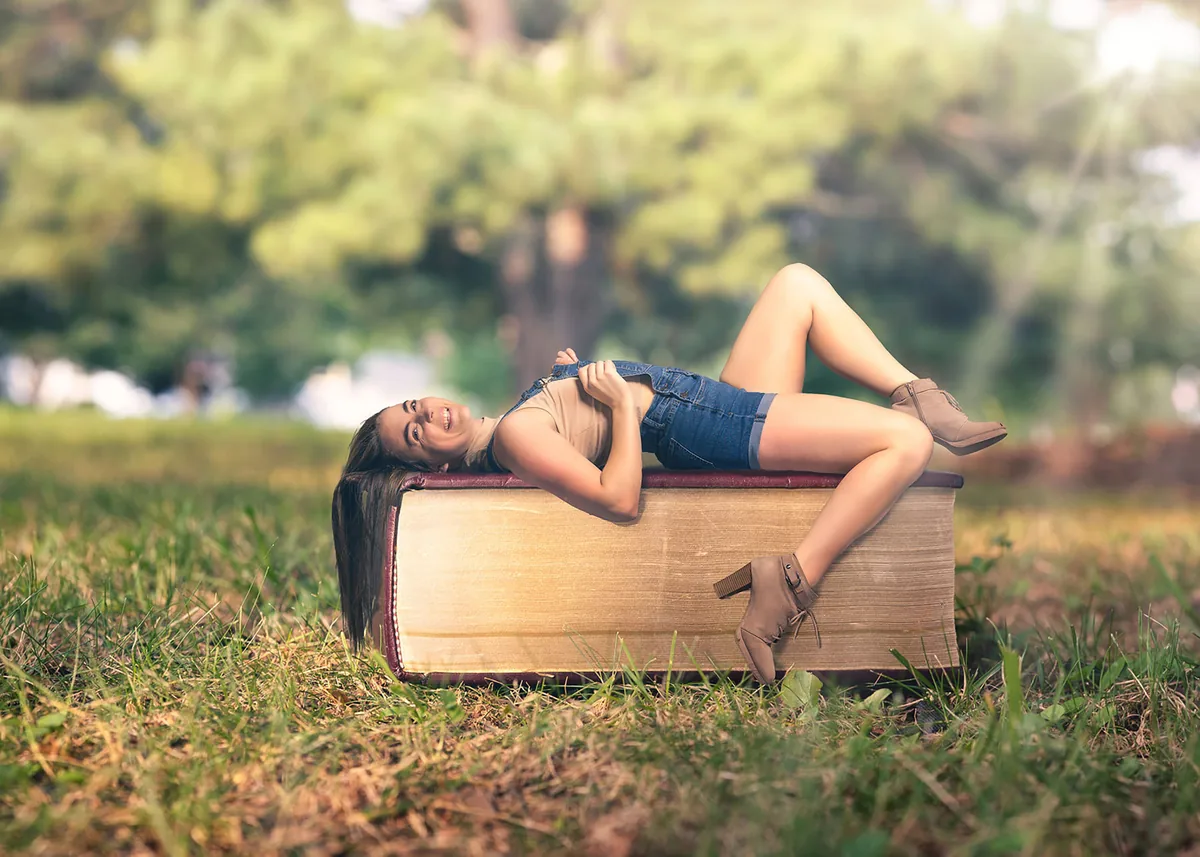 A young woman lies on her back across an oversized book resting on grass, wearing a beige sleeveless top, denim shorts, and taupe ankle boots in soft daylight.