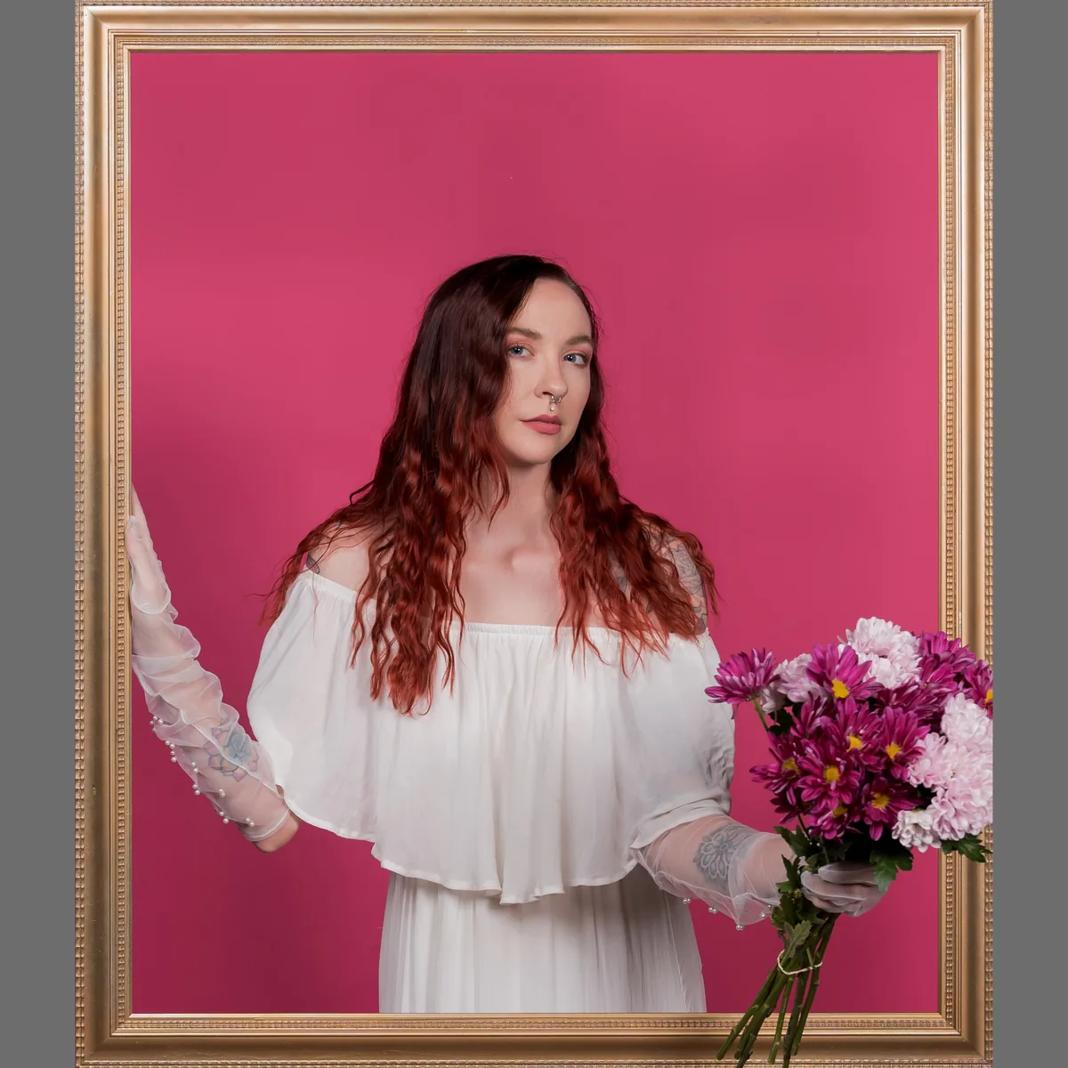 A woman with long wavy red hair stands holding a bouquet of purple and pink flowers, framed by a large gold picture frame, against a solid bright pink background.