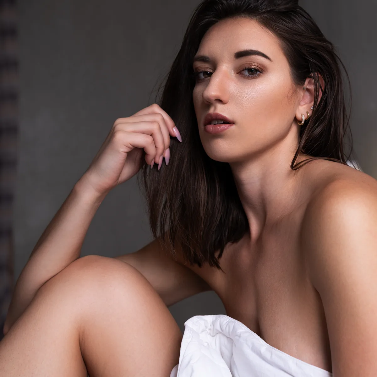 A young woman with shoulder-length dark hair sits with her knee drawn up, her bare shoulder and upper chest visible beneath a white fabric, illuminated by soft, natural light.
