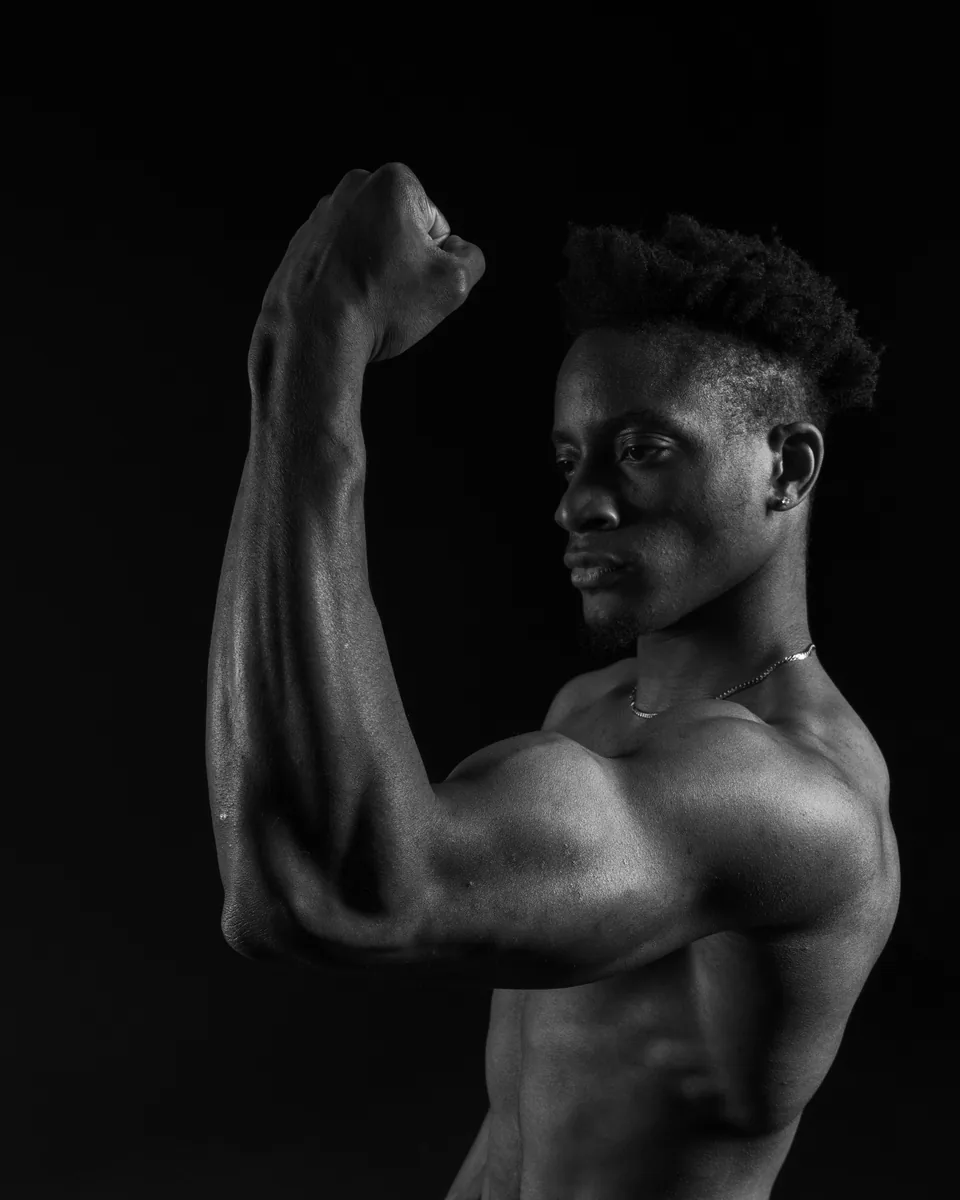 Black and white image of a shirtless man shown in profile with his right arm raised and fist clenched, highlighting muscle definition against a dark background.