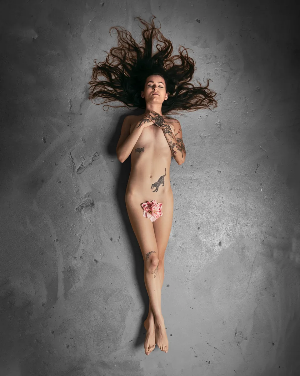 A nude person with long dark hair lies on a textured grey surface, hands clasped at chest, tattoos visible on arms and torso, with a flower placed over the pelvic area.