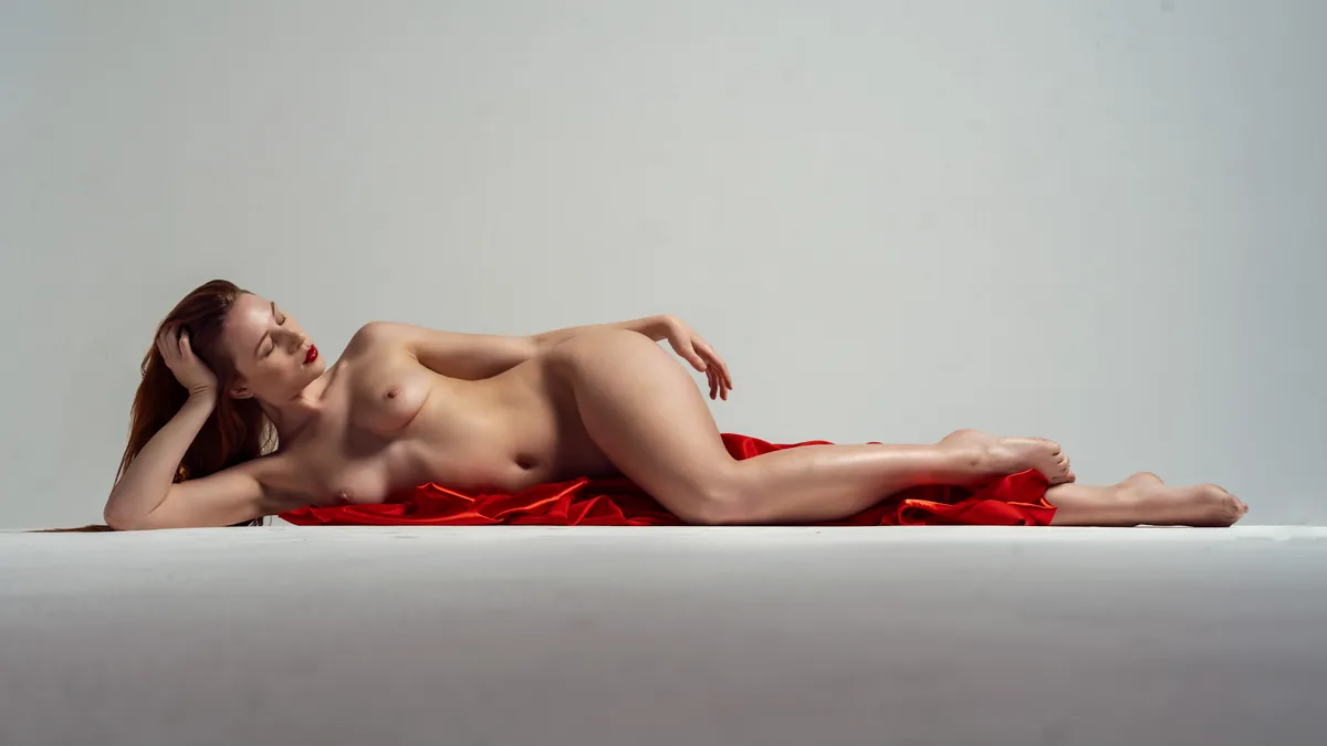 A nude woman with red hair lies on her side on a grey surface with red fabric beneath her, lit softly from above, her eyes closed and hand resting on her head.