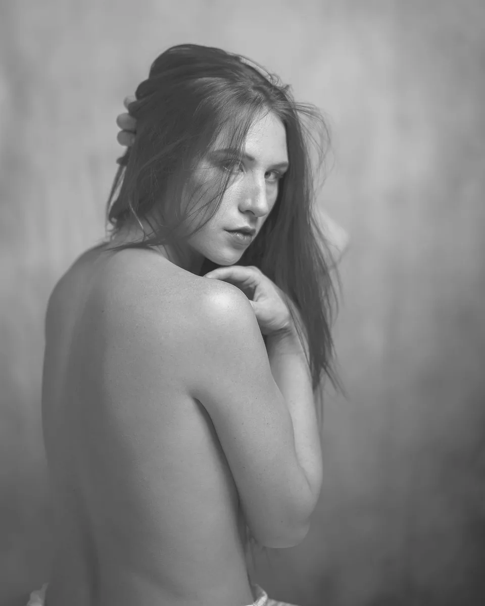 Black and white image of a bare-backed person with long hair looking over their shoulder, one hand touching their neck and the other in their hair, against a soft-focus background.