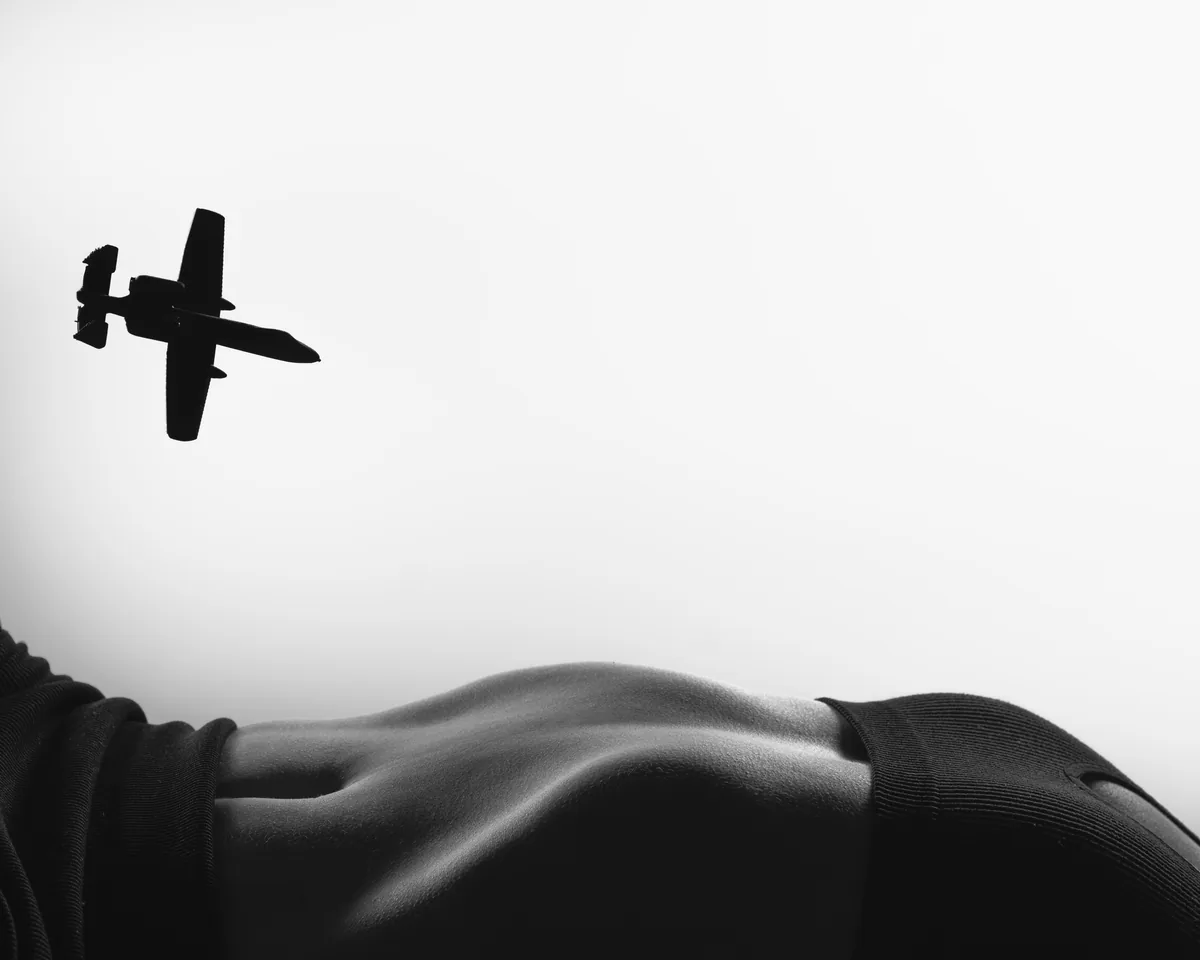 Black and white image showing a close-up of a curved human torso in dark fabric with a silhouette of an airplane flying overhead against a bright background.