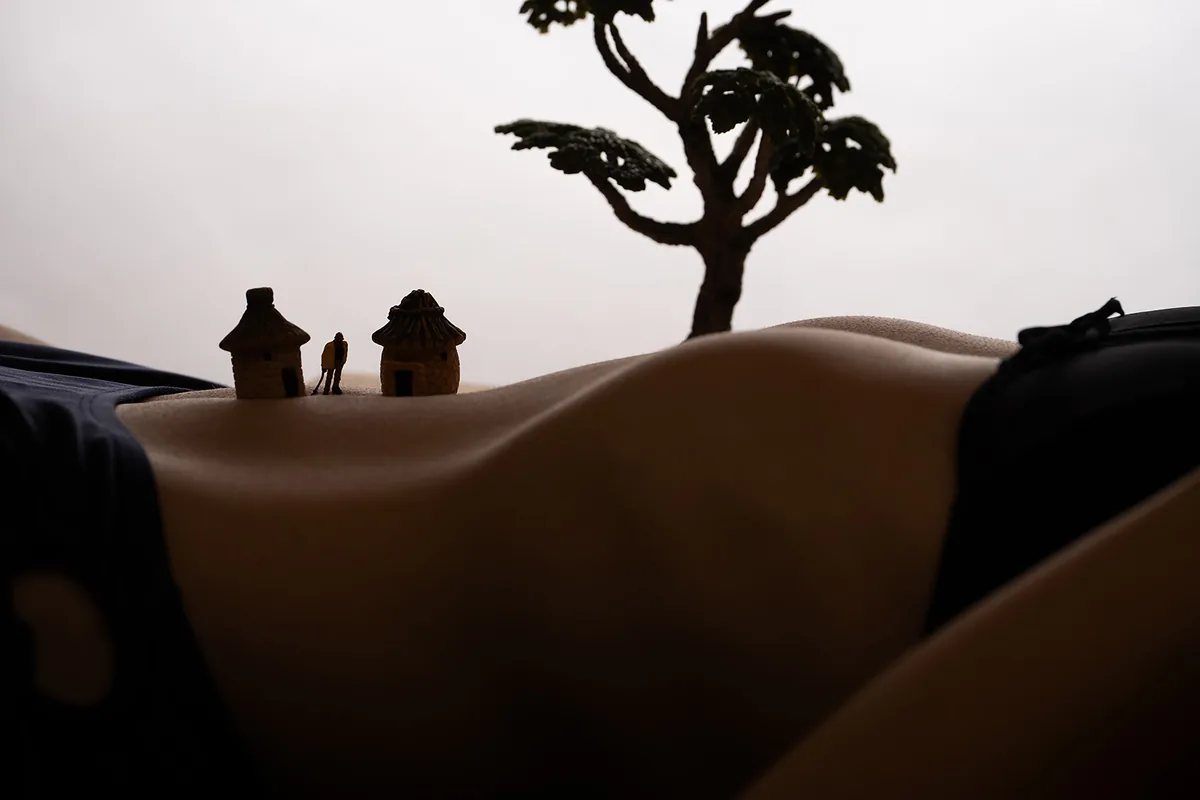A low-lit image showing a torso in black clothing, with miniature huts, a small figure, and a tree placed on the skin’s curves, creating a scaled landscape effect.