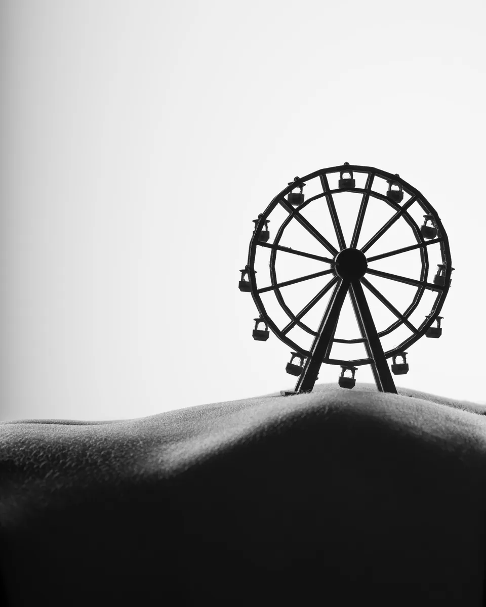 Black and white image showing a textured, curved surface in the foreground resembling a hill, with a small Ferris wheel structure placed behind the curve against a bright sky.