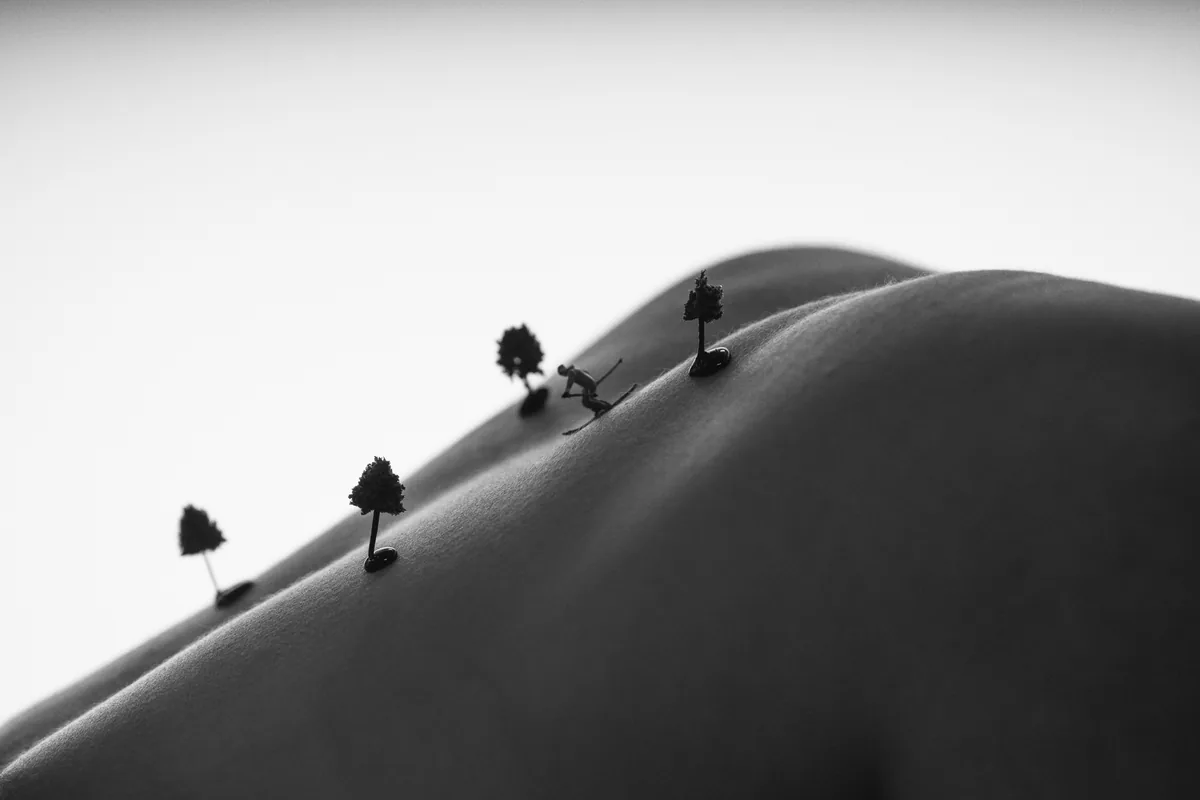 Black and white image of a close-up human back curved like hills with small model trees and a miniature skier placed on the skin, lit softly from the side.
