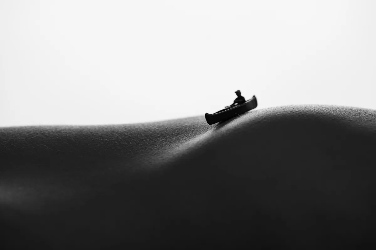 Black and white image showing a miniature figure in a canoe positioned on a curved section of human skin, lit softly to emphasize texture and shape.