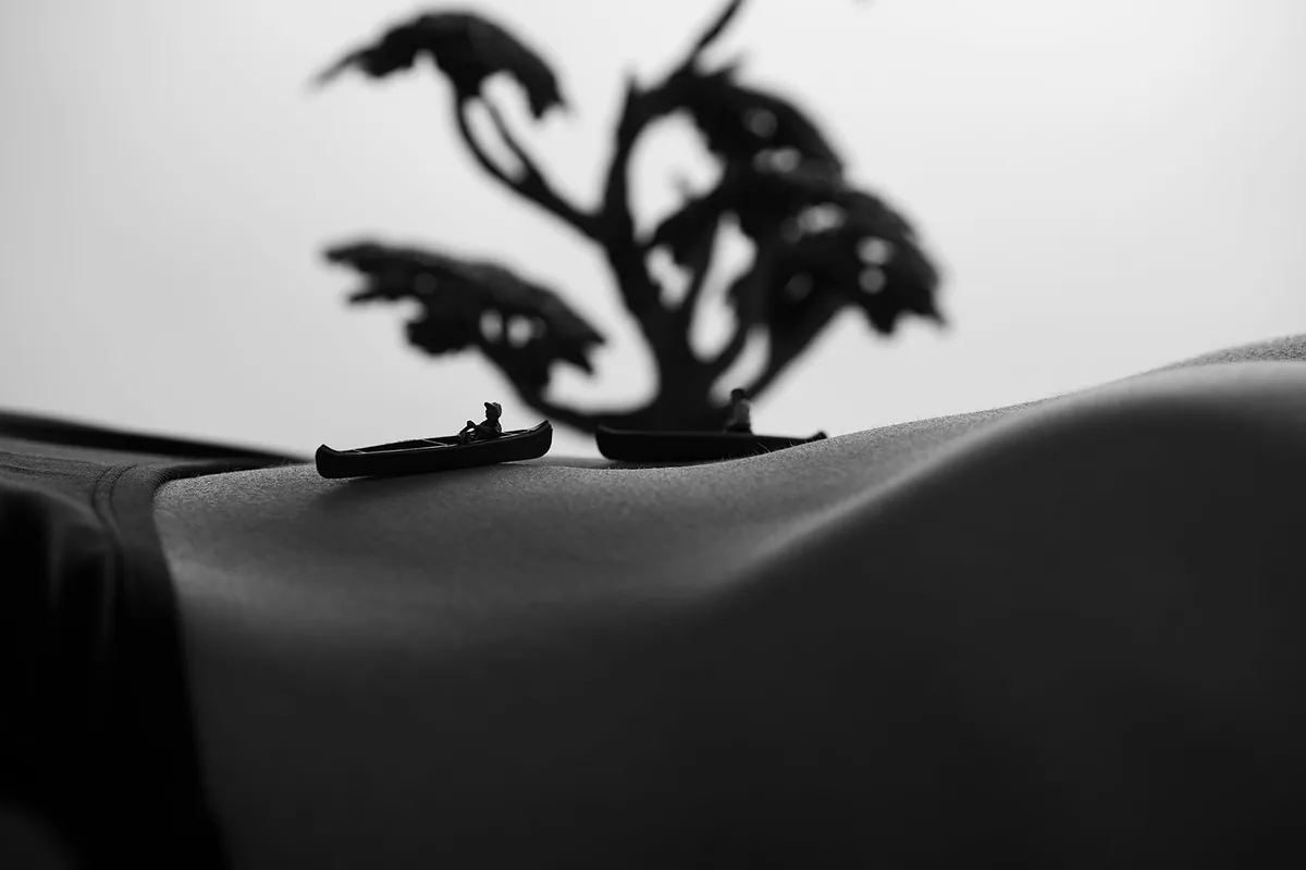 Black and white close-up of a curved section of skin with two small canoes and miniature figures placed on the surface, with a blurred tree-shaped object in the background.