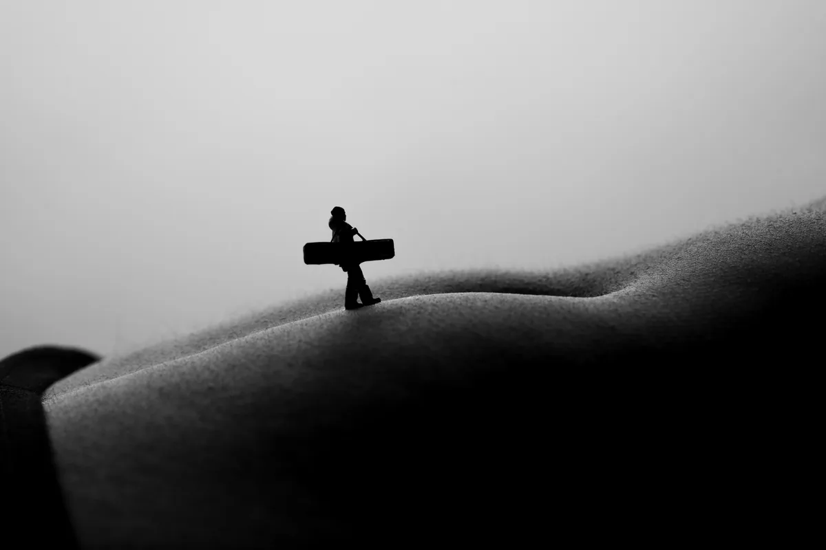 A small figurine carrying a rectangular object walks across the textured, curved surface of a body under soft, diffused light creating a silhouette effect.
