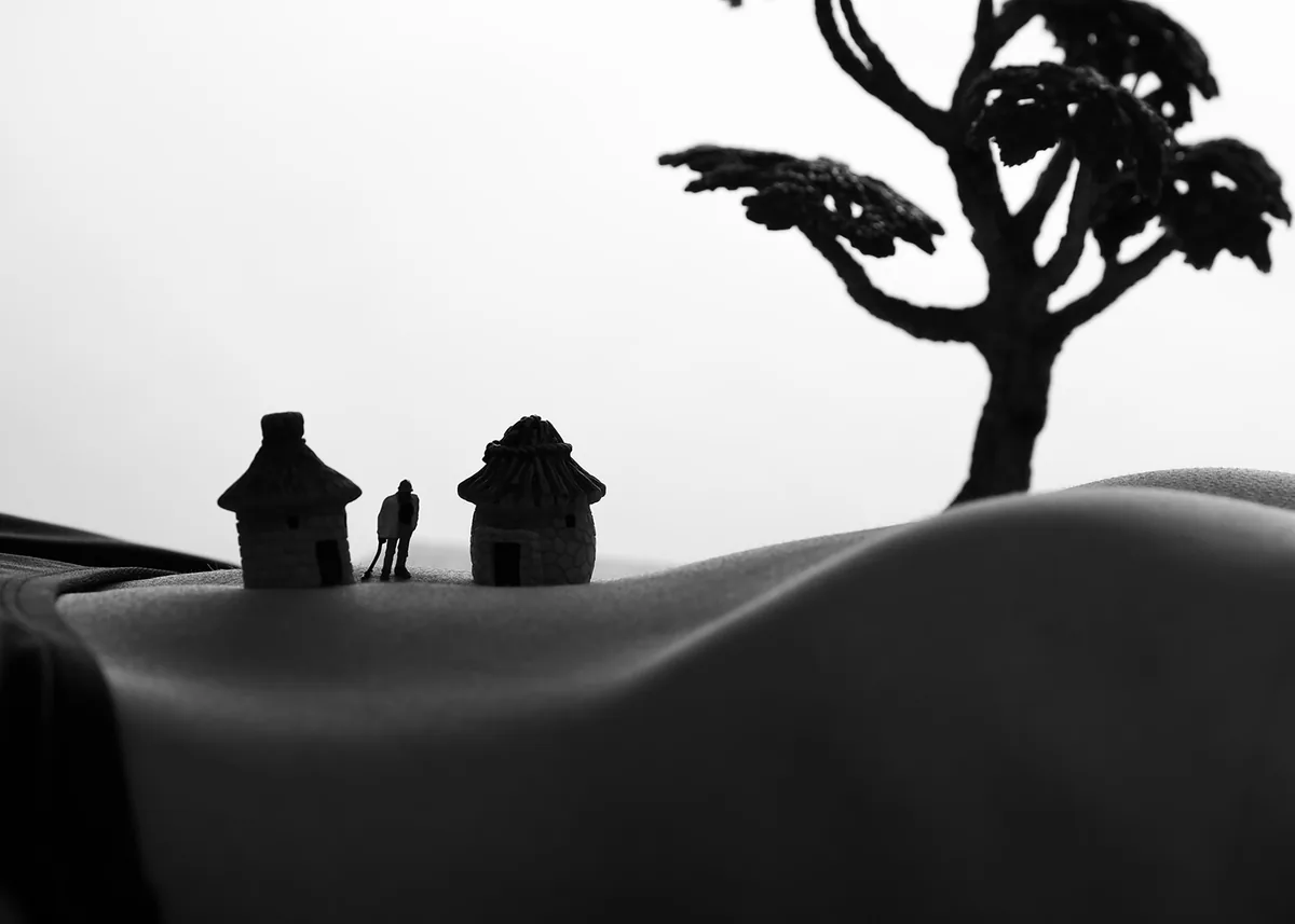 Black and white image showing two small model huts, a figure, and a tree placed on a smooth, curved section of a person's torso resembling a landscape with soft lighting and gentle shadows.