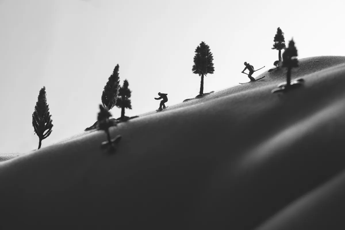 Black and white image showing miniature skier figures moving down a smooth curved surface resembling a snowy hill, dotted with small model trees simulating a winter landscape.