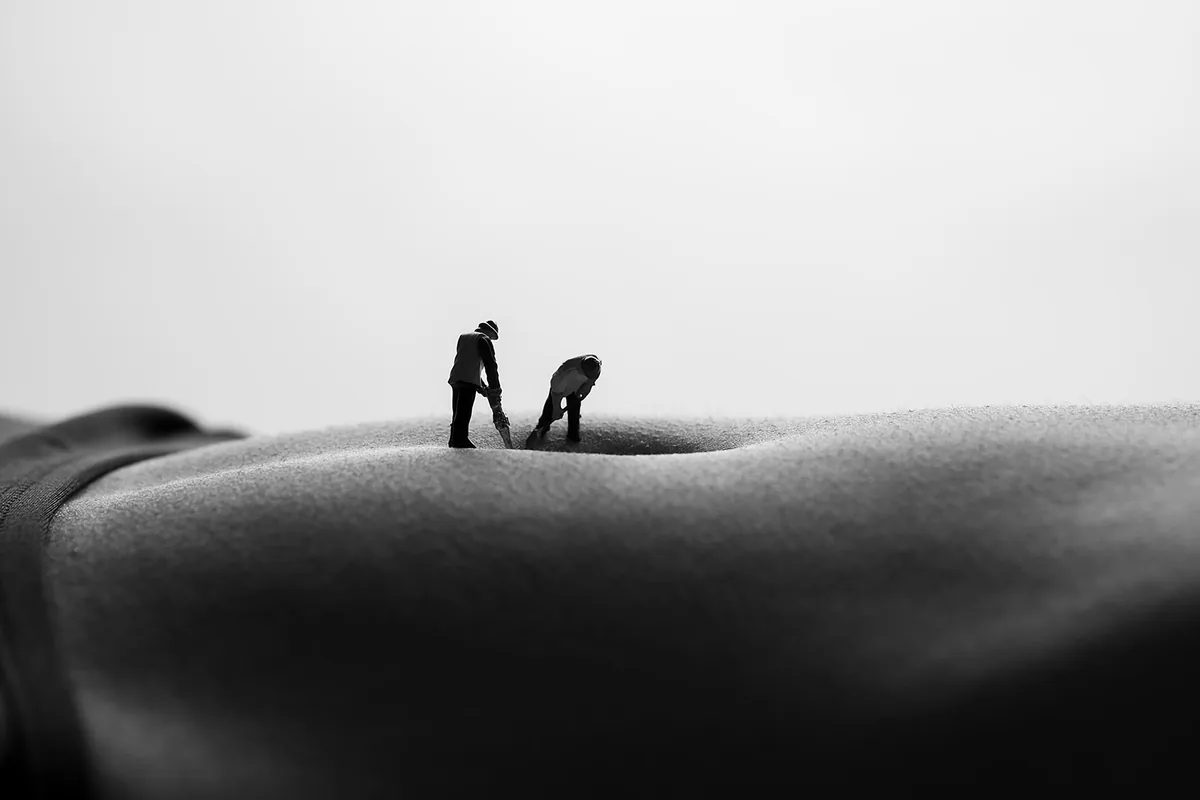 Black and white image of a close-up torso with two tiny worker figures positioned near a belly button, creating an illusion of a vast landscape under diffused light.