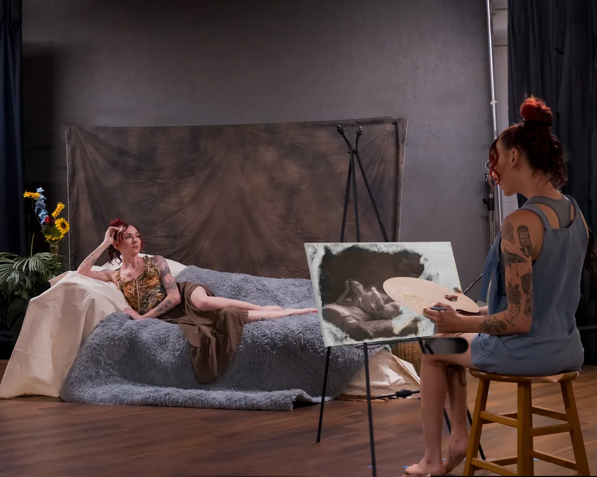 A woman with tattoos sits barefoot on a wooden stool painting a reclining version of herself on a textured canvas, lit softly in a studio with muted tones and fabric draped background.