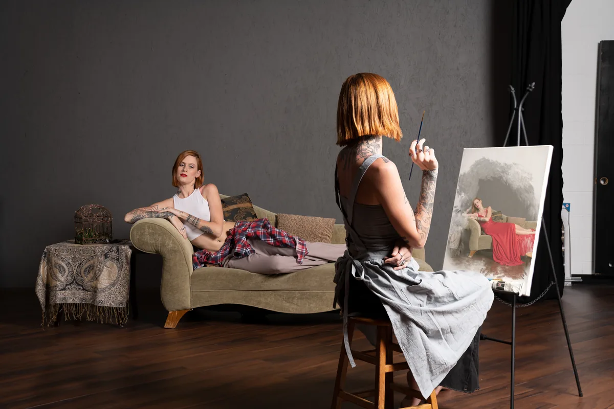 A tattooed woman with orange bob hair sits on a stool painting a canvas that depicts the same woman reclining on a couch in a red dress, while the real woman wears casual clothing in softer light.