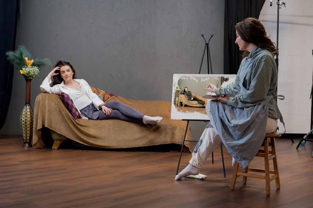 A woman in casual clothing sits on a wooden stool painting a portrait of herself reclining on a couch, with soft lighting in a studio setting.