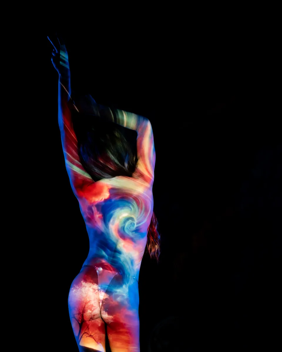 A person shown from behind with arms raised and crossed above the head, their back illuminated by projection of swirling colors and sky imagery on bare skin against a dark background.
