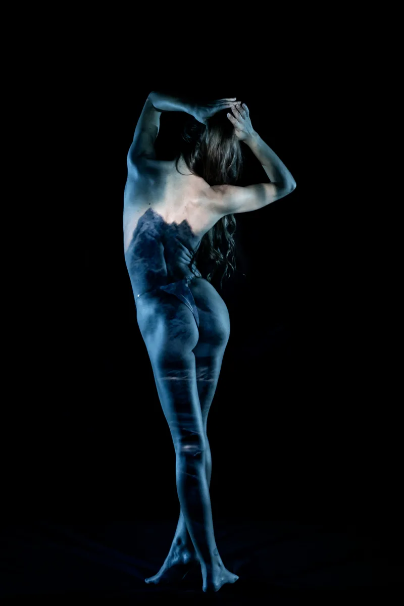 A person stands barefoot with their back to the camera, arms raised holding their head, while a blue-toned mountain landscape is projected across their body against a dark background.