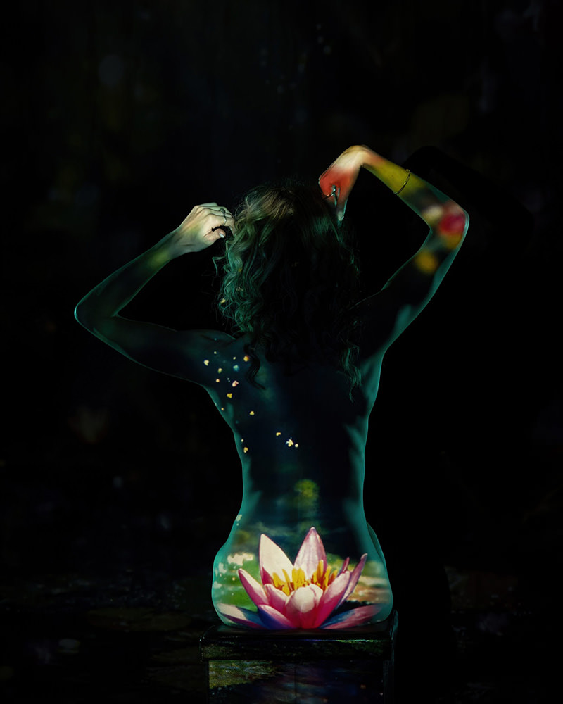 Nude person sitting with back facing camera, illuminated by a colorful projection of a pink and yellow water lily and small scattered lights against a dark background