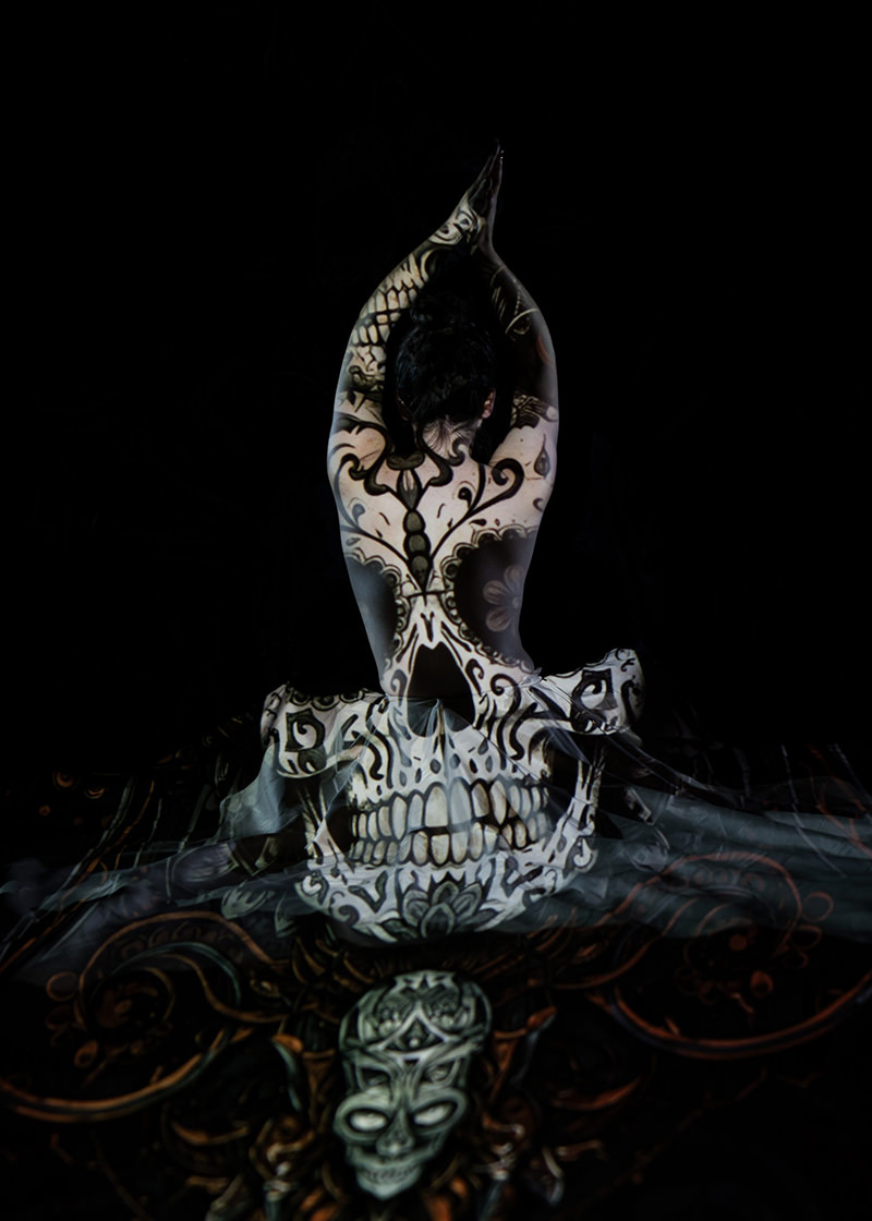 Person seated in a meditative pose with arms raised, their back illuminated by an intricate black and white skull design projected onto their skin and surrounding fabric against a dark background