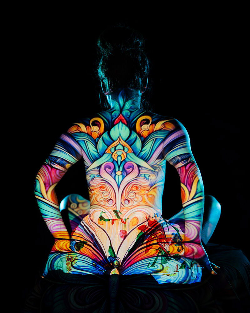 Back of a seated person with intricate, brightly colored patterns projected across their skin, set against a black background