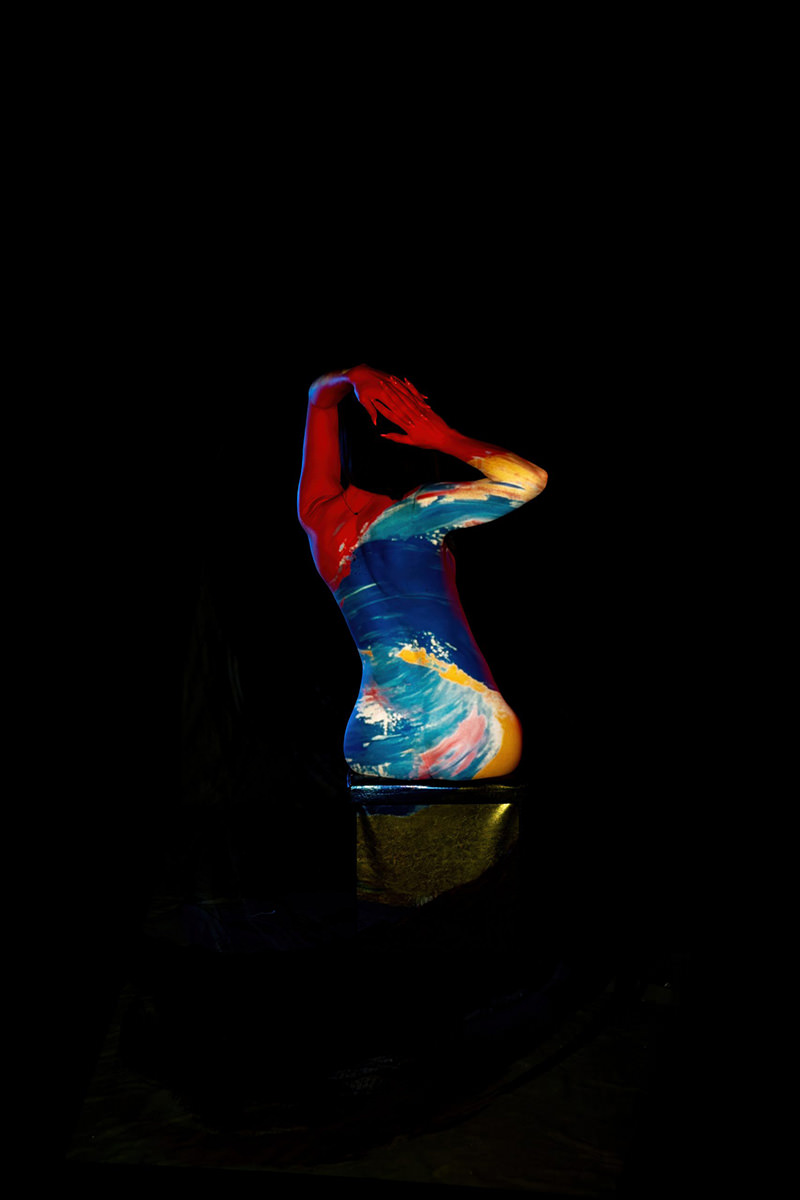 Torso and arms painted with bright red, blue, yellow, and white abstract strokes, posed against a dark background with hands raised near the head, creating a striking artistic effect