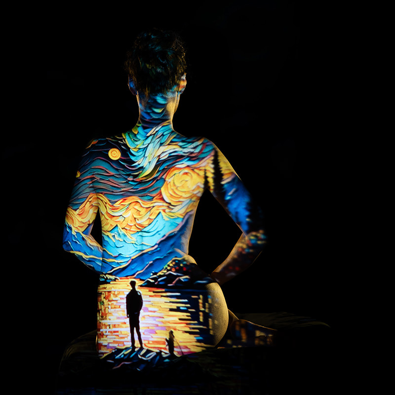 Back view of a person with a colorful projection of a starry night sky and a figure standing by the water, casting vivid yellows, blues, and oranges across their skin against a black background