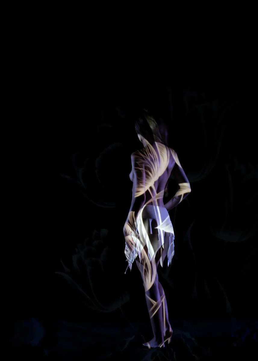 A woman stands barefoot and turned slightly away with a projection of light patterns resembling leaves cast across her bare back and white fabric skirt in a dark studio.