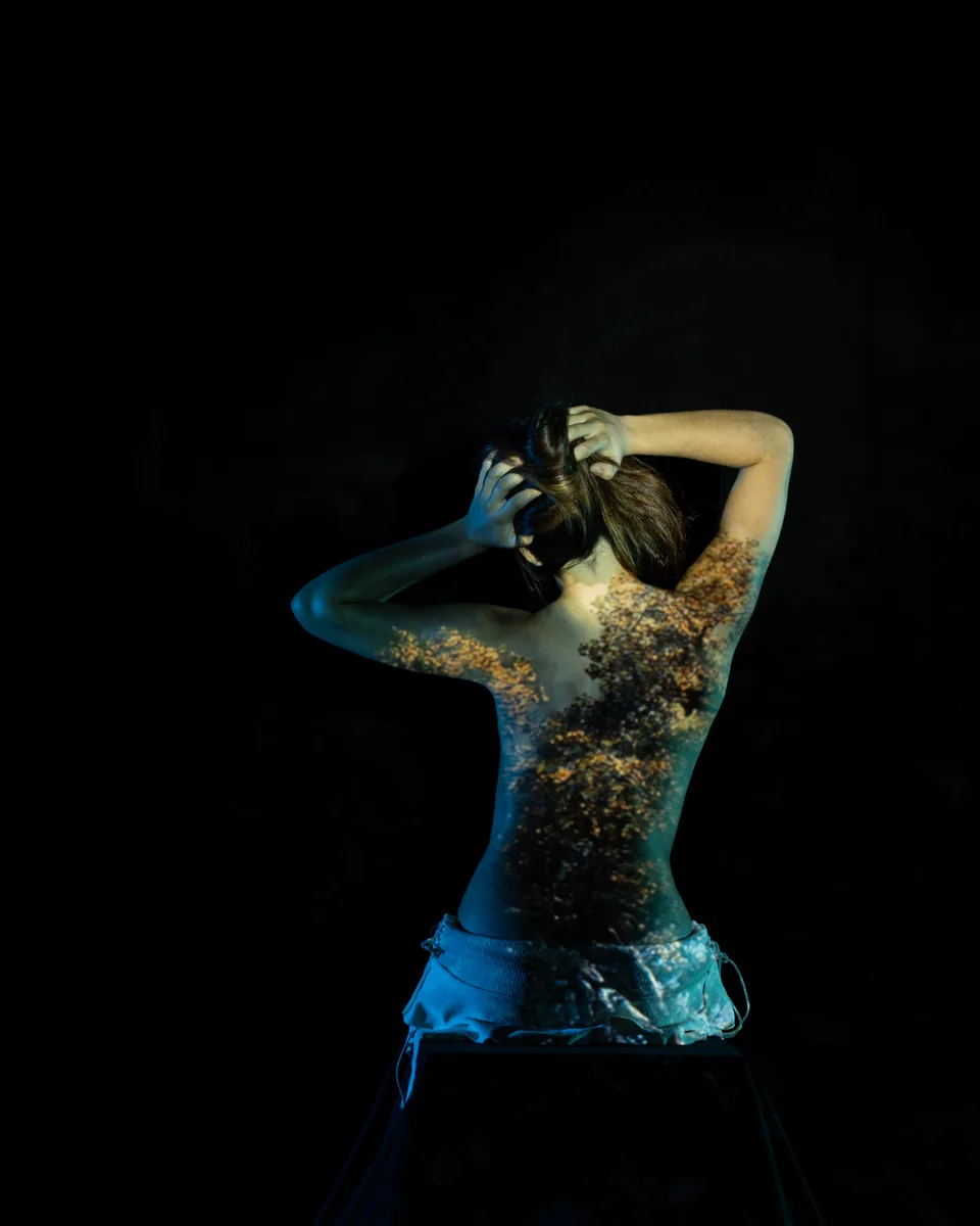 A seated person with their back to the camera, arms raised holding their hair, with a projection of tree branches and leaves cast across their back and shoulders in a dark studio.