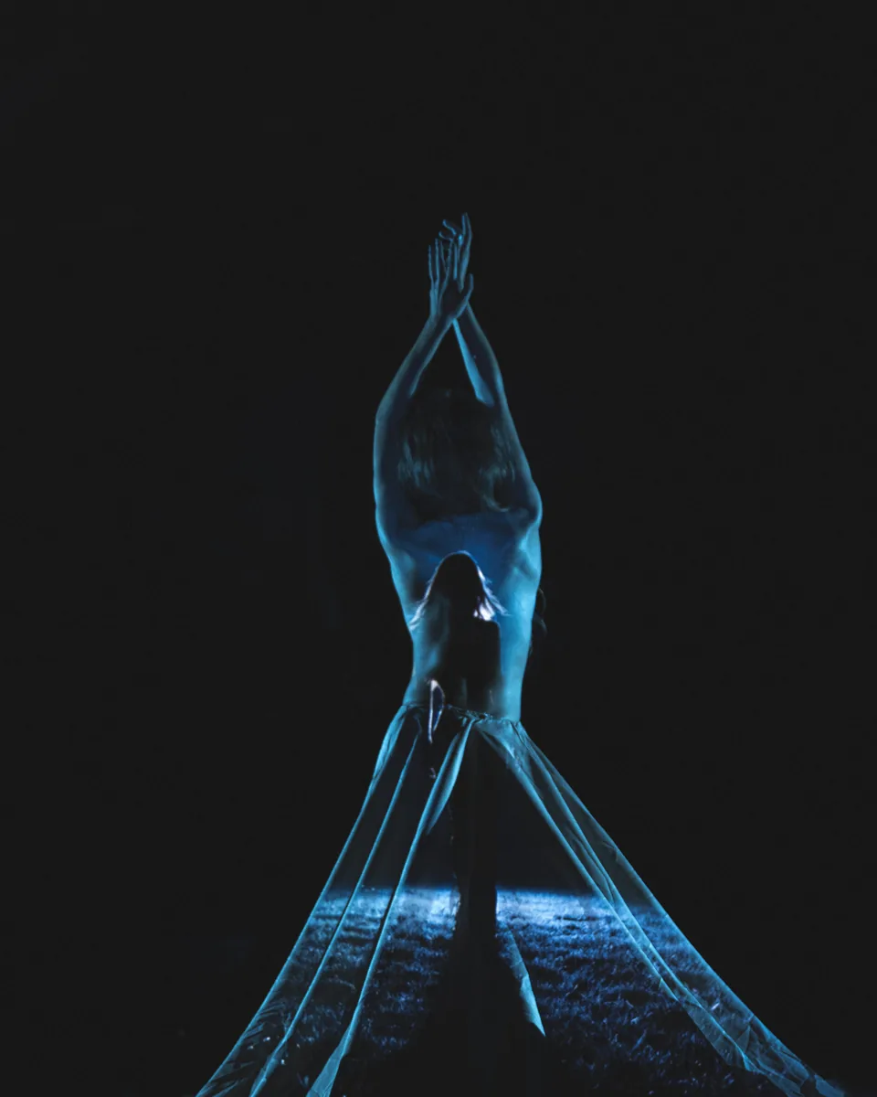 A person stands with arms raised above their head, back facing the viewer, with blue light projected onto their bare skin and semi-transparent fabric draping downward, set against a dark background.