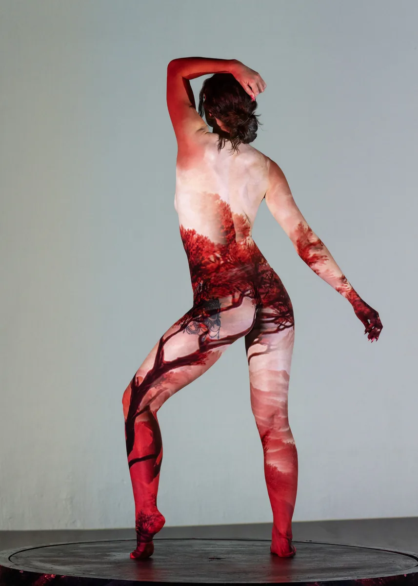 A person stands on a circular platform with a red landscape projection covering their back and legs, one arm raised behind their head and the other extended sideways against a light gray background.