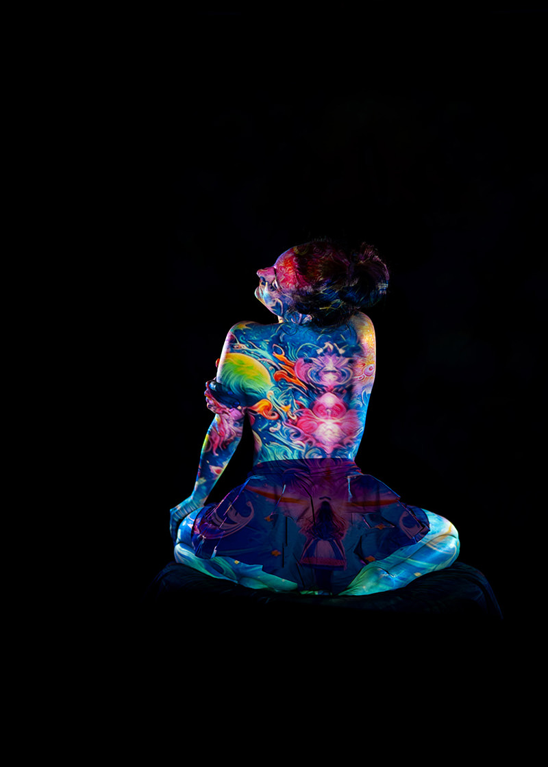 Woman sitting cross-legged with her back facing the camera, her bare upper body covered in vibrant, swirling cosmic and floral body paint patterns against a black background