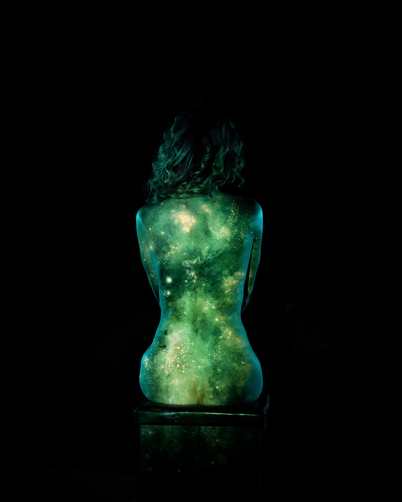 A person sitting on a box with their back facing the viewer, with a green and yellow galaxy projection covering their entire back against a black background