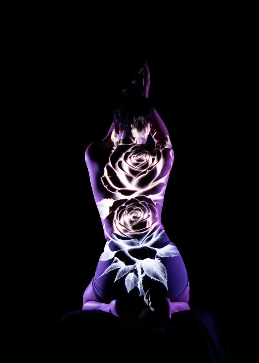 Person kneeling with arms raised above head, back facing viewer, illuminated by projected image of large white roses and leaves against dark background