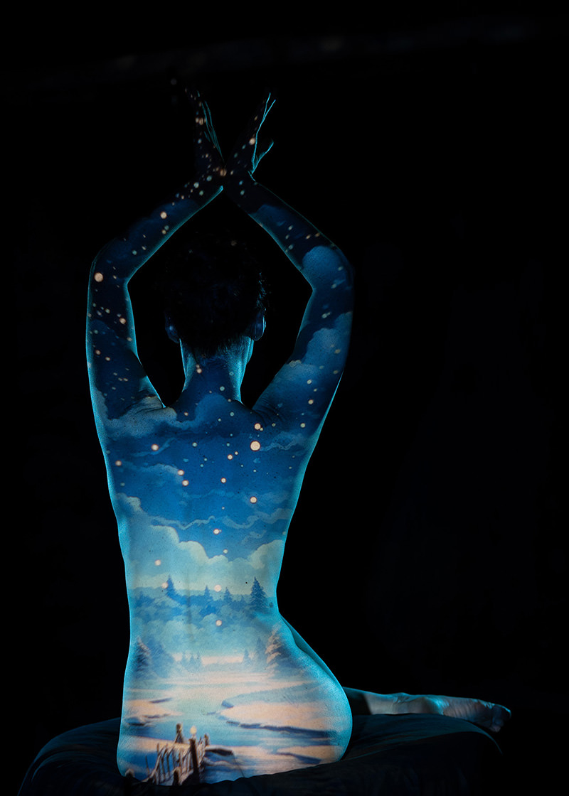 Back view of a seated person with arms raised and crossed above head, their bare skin illuminated by a projection of a starry night sky and a serene landscape with trees and a river.
