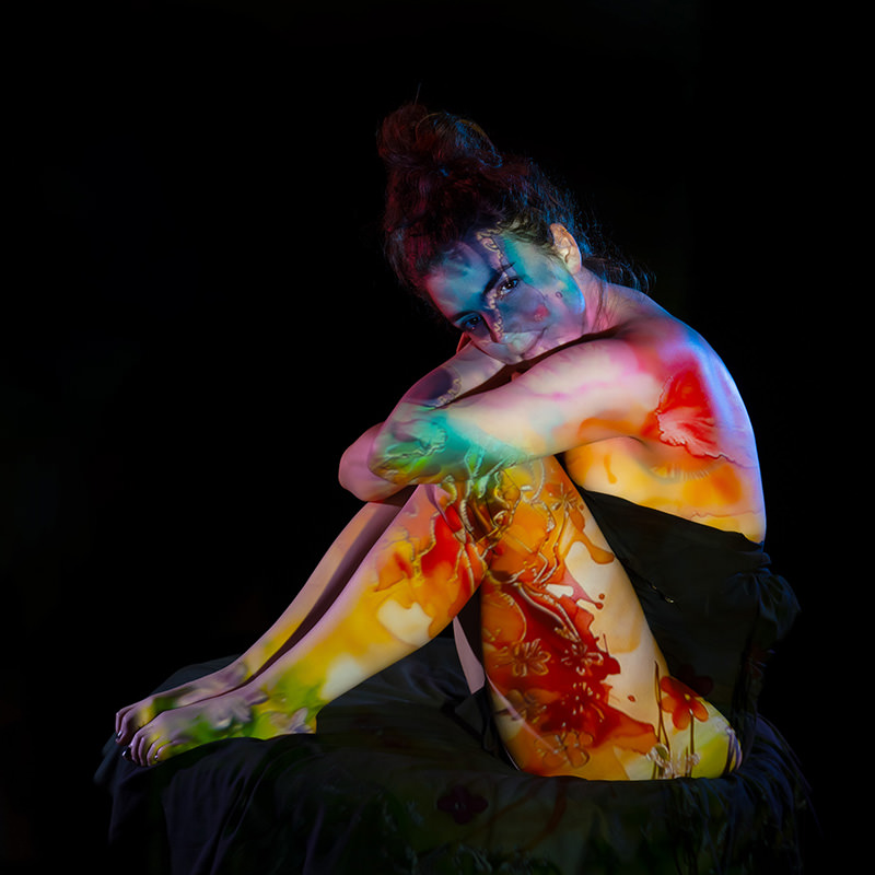 A woman sits with knees drawn close to her chest, her body illuminated by vibrant projections of abstract floral patterns in red, yellow, green, and blue hues against a black background.