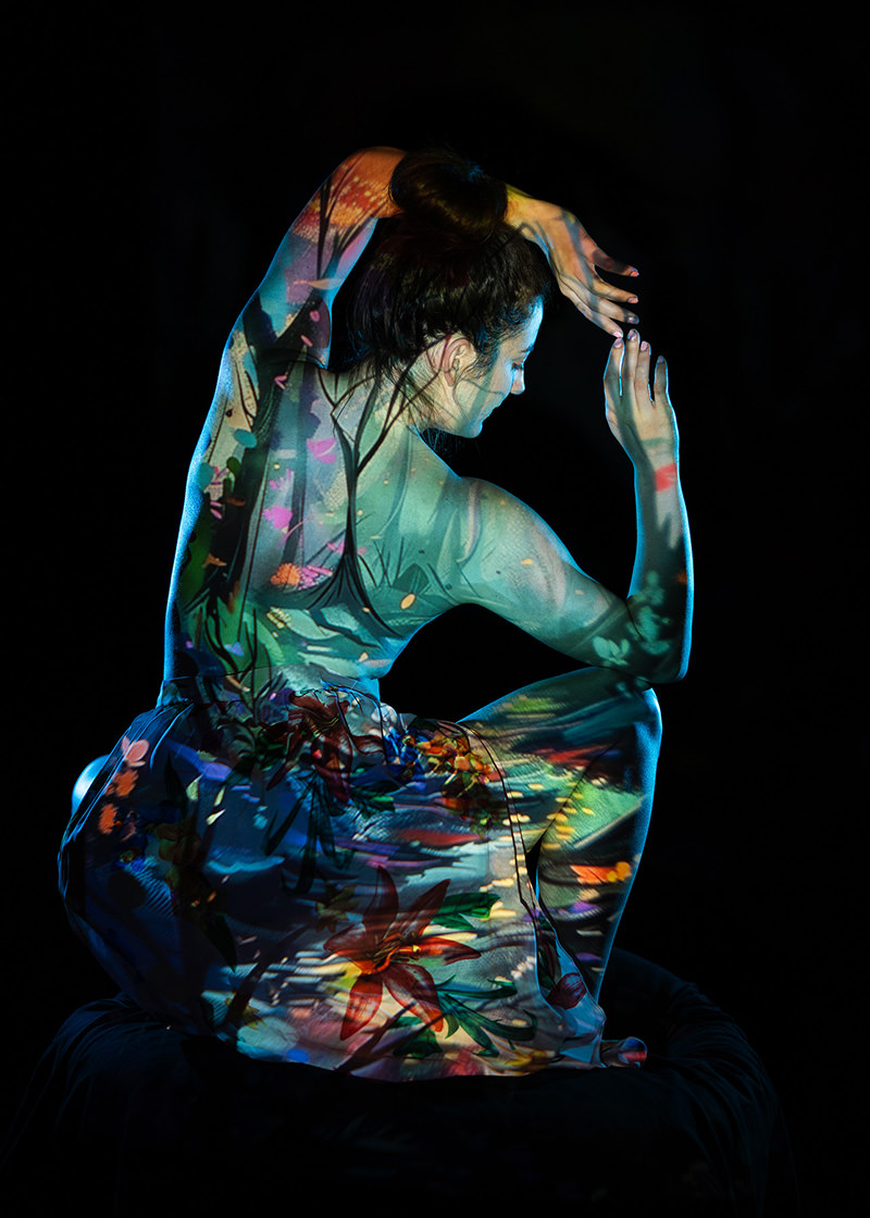A woman with hair in a bun poses with arms curved above her head, sitting against a black background. A colorful nature scene with trees, flowers, and leaves is projected onto her body and clothing.