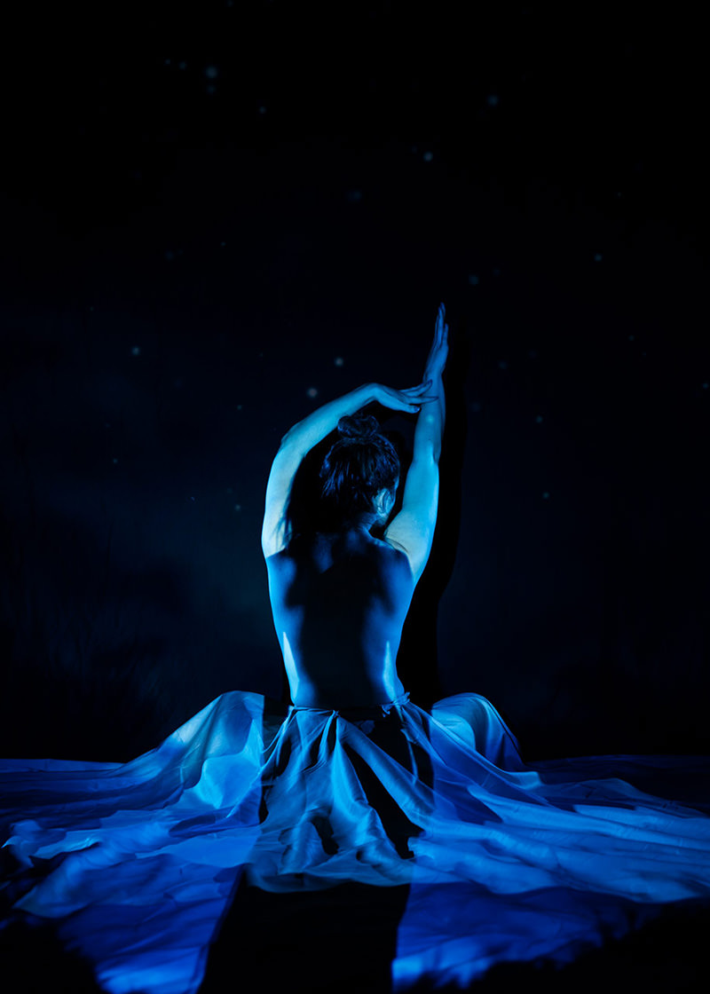Bareback person seated on the floor with arms raised and curved overhead, wearing a flowing blue skirt, illuminated by blue lighting against a dark backdrop.