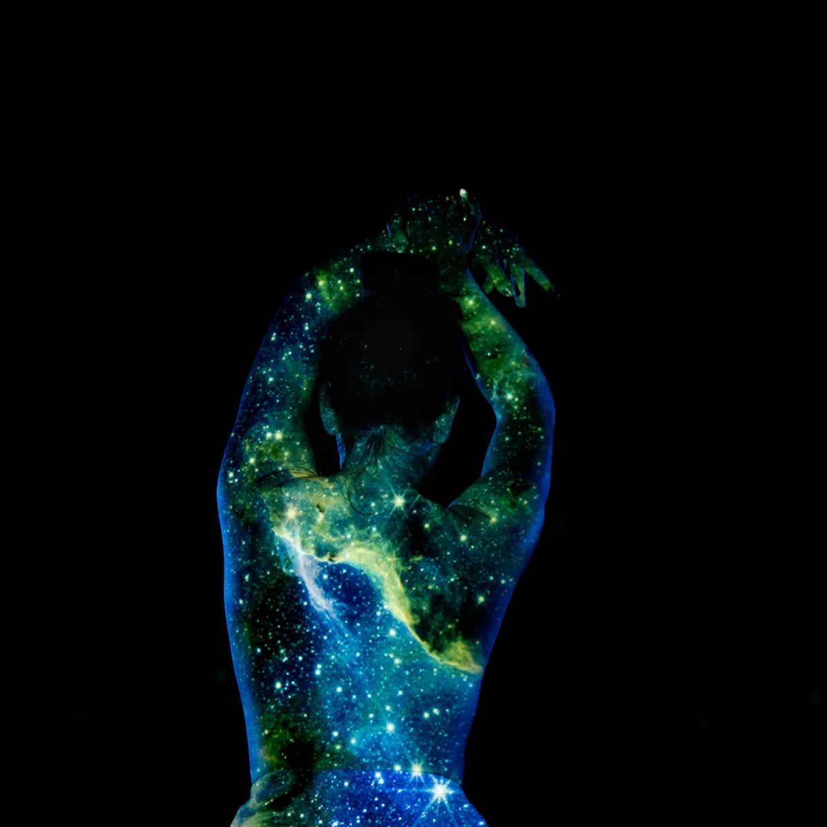 A figure viewed from behind with arms raised above the head, covered in a projected pattern of stars and nebulae in blue, green, and yellow hues against a dark background.