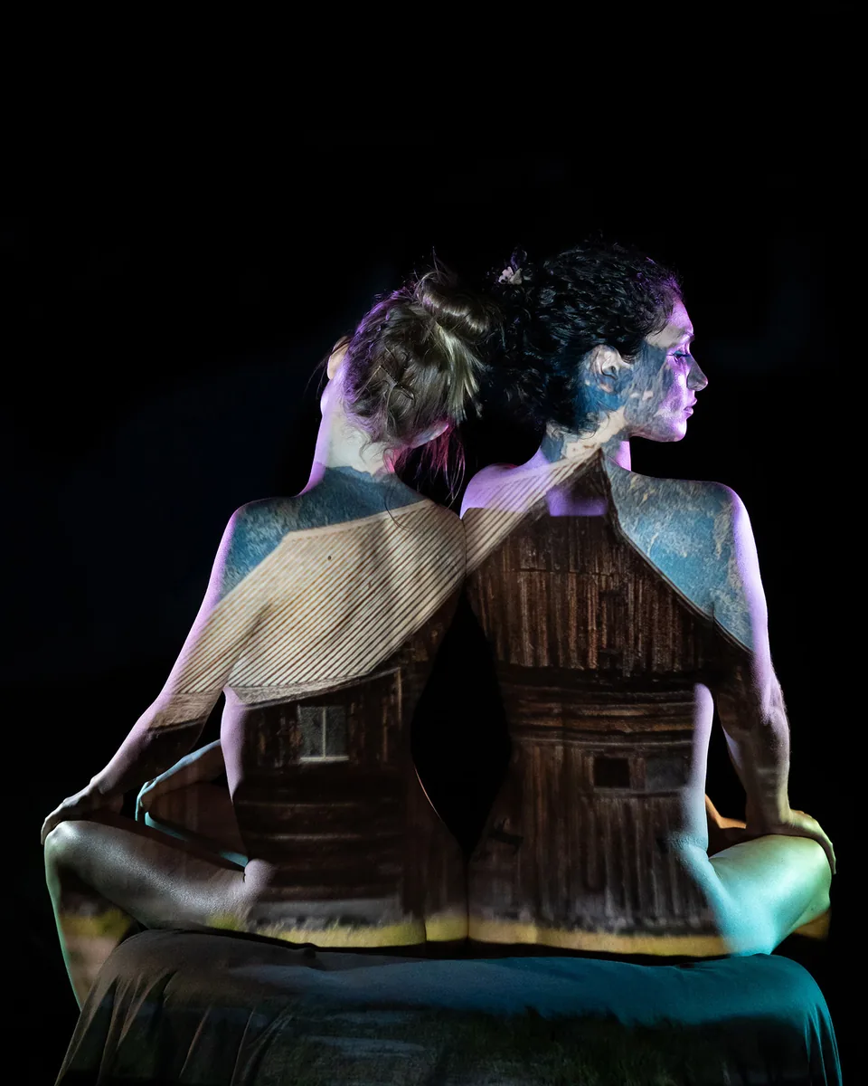 Two seated figures with backs to the camera, light and artwork of a rustic barn projected across their skin, creating a textured layered effect against a black background.