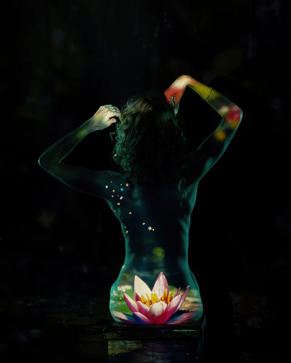 A figure sitting with their back to the camera, with soft light and a water lily projected across the lower back and arm, creating a quiet, contemplative mood.