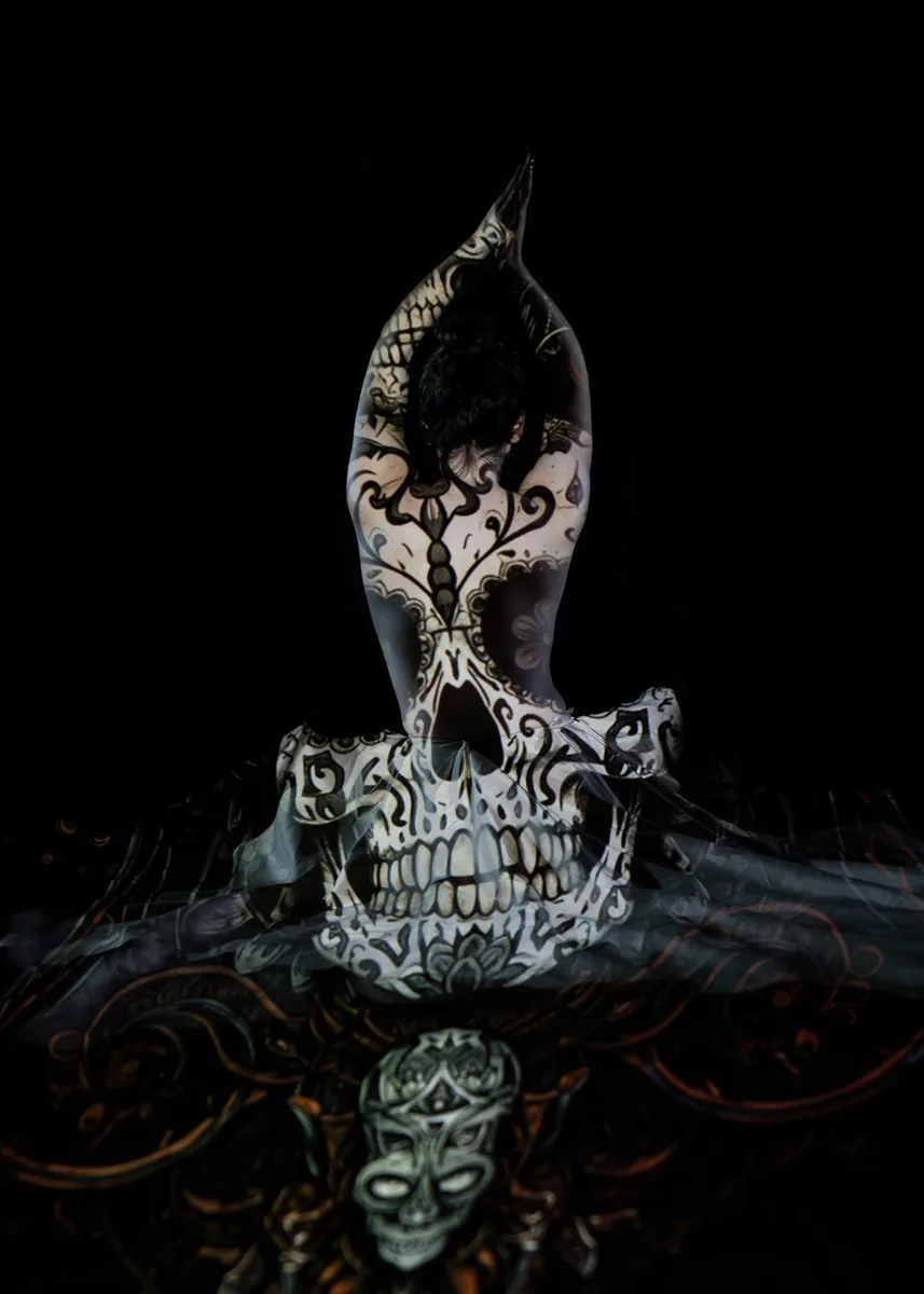 A person sits with their back to the camera, arms raised and pressed together above their head, while black and white skull and ornamental patterns are projected onto their skin and a sheer fabric surrounding them, set against a dark background.