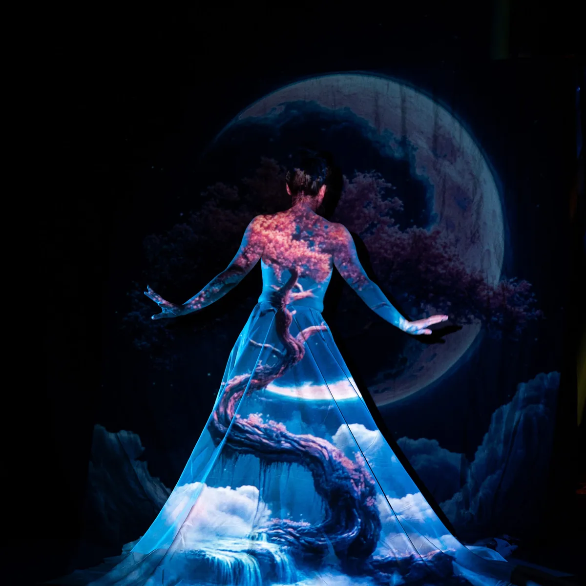 A person faces away with a soft blue light projecting a twisting tree with pink blossoms and clouds across their back and a flowing skirt, set against a large moon backdrop in darkness.