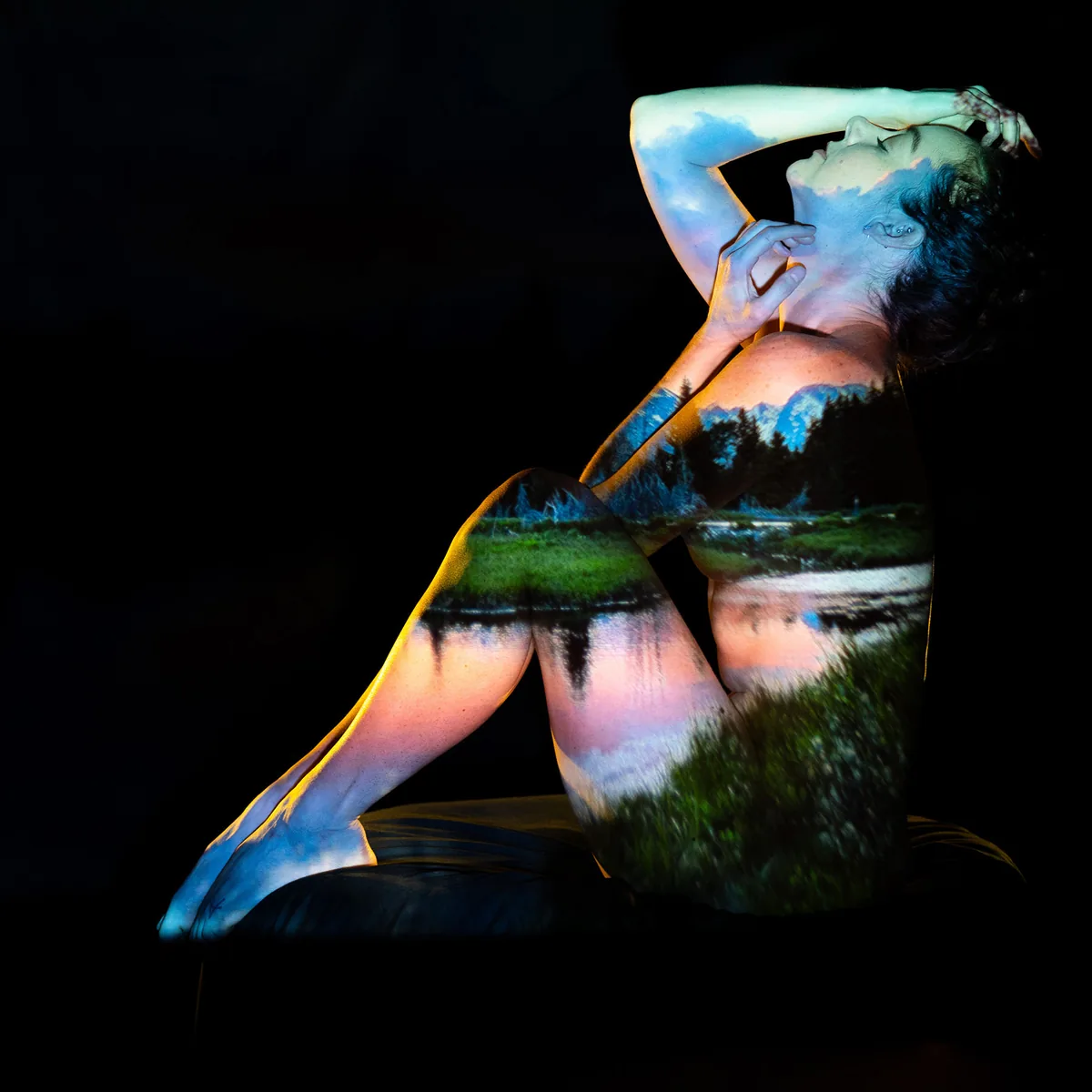 A seated figure with a landscape projection of trees, water, and sky on their skin, posed with one hand on their head and the other near their face, set against a black background.