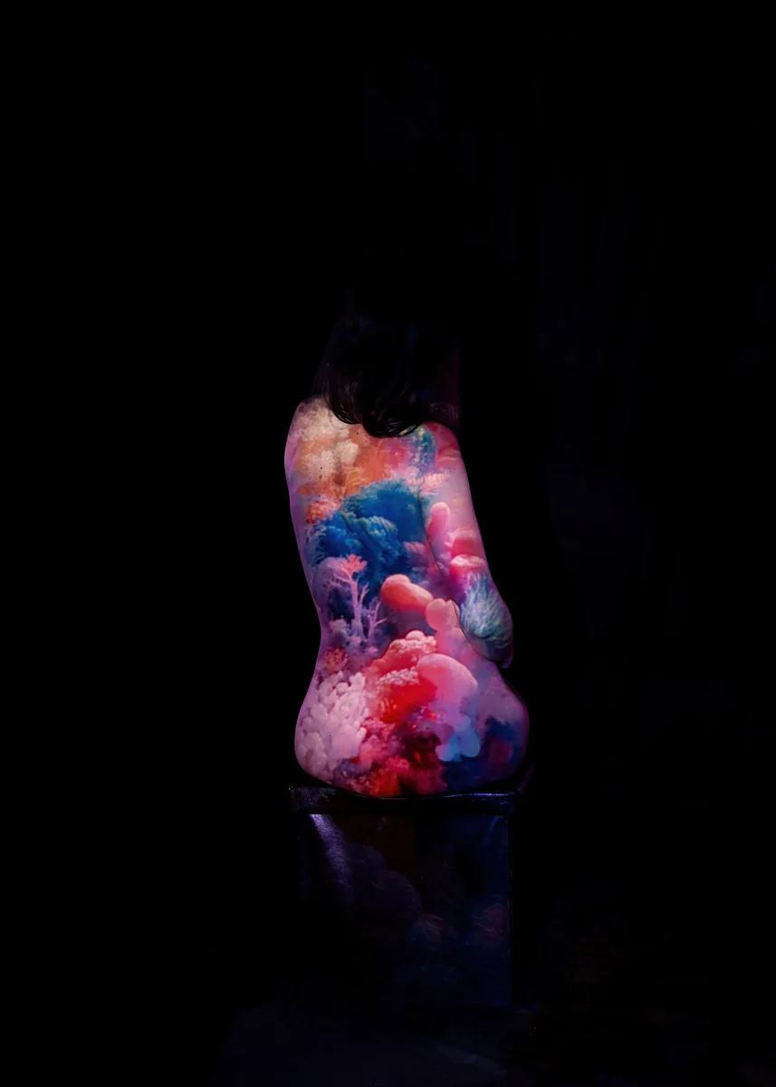 A seated figure with back turned, illuminated by soft, colorful projections resembling abstract underwater coral forms in pink, blue, and orange hues, against a dark background.