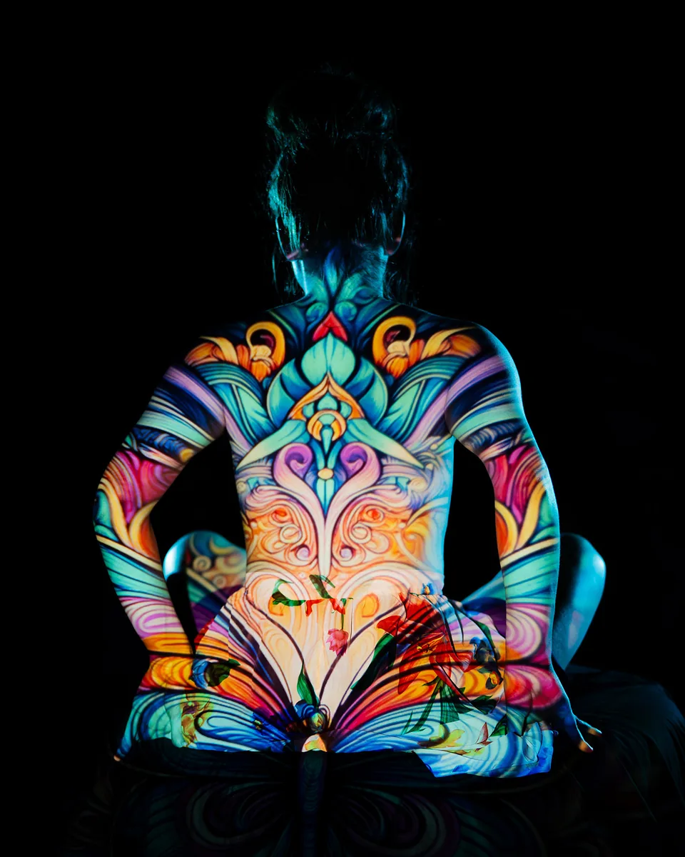 A person sits with their back to the camera, illuminated by colorful, detailed projection art featuring symmetrical, swirling designs in shades of blue, orange, and purple against a dark background.