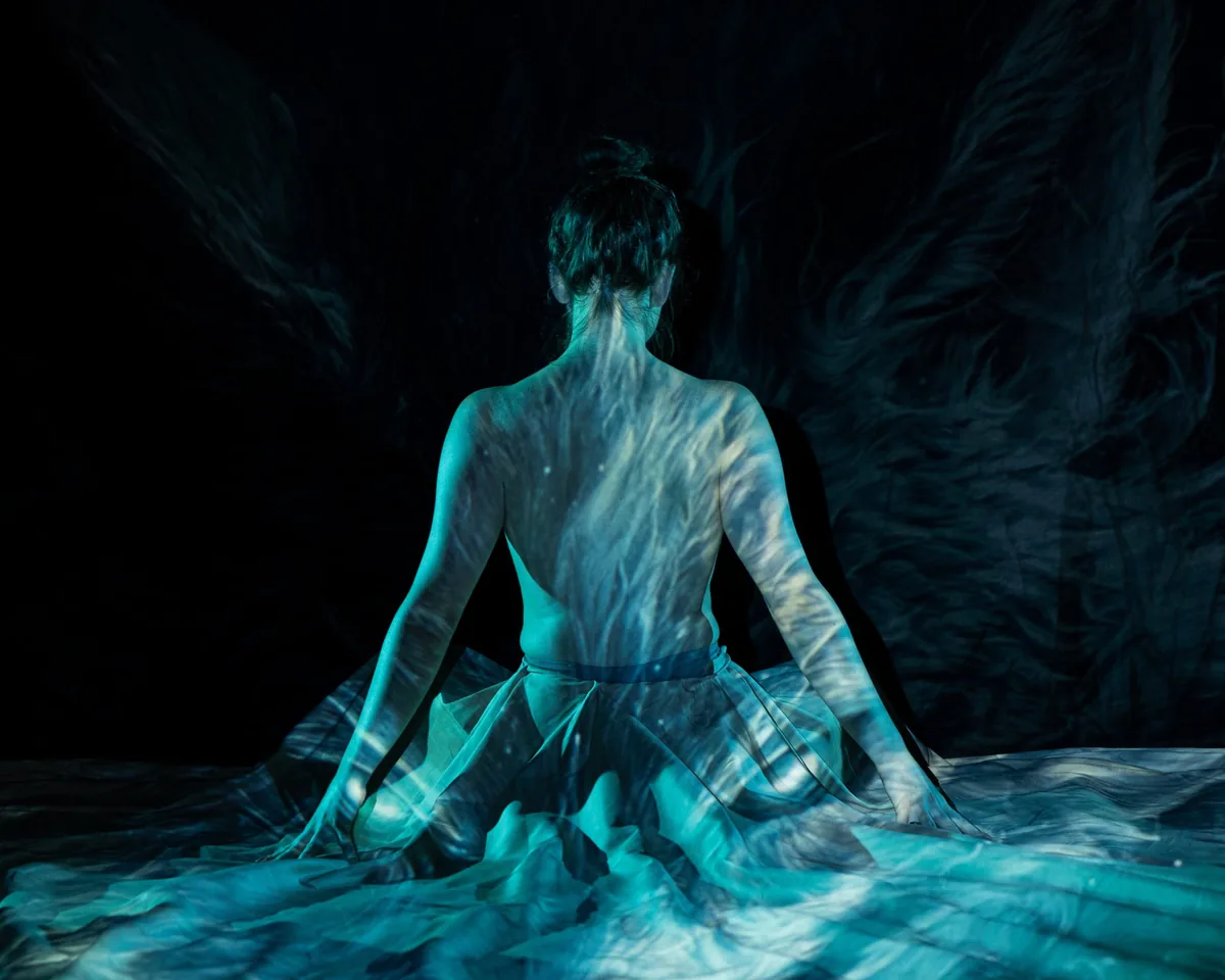 A person seated with their back facing the camera, bare upper body illuminated by blue-toned flowing projection patterns, wearing a sheer fabric skirt, evoking a calm and contemplative atmosphere.